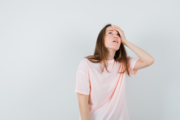 Young female holding hand on forehead in pink t-shirt and looking wistful. front view.