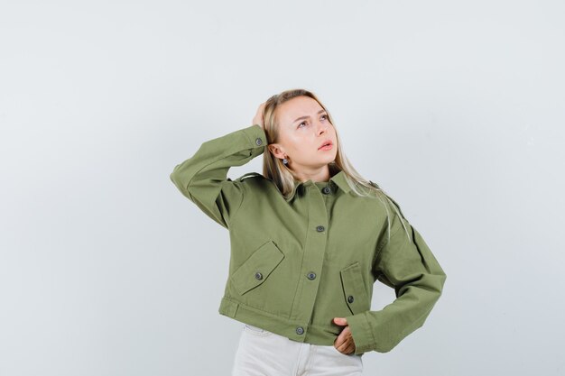 Young female holding hand on head in green jacket,jeans and looking focused , front view.