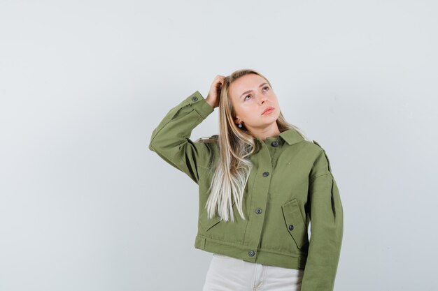 Young female holding hand on head while posing in green jacket,jeans and looking thoughtful. front view.