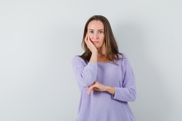 Young female holding hand on her cheek in lilac blouse and looking calm 