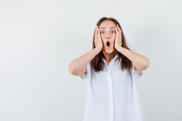 Young female holding hand on her face in white blouse and looking frightened.