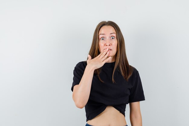 Young female holding hand on mouth in black blouse and looking scared