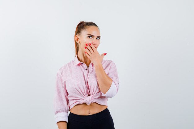 Young female holding hand on mouth in casual shirt, pants and looking puzzled