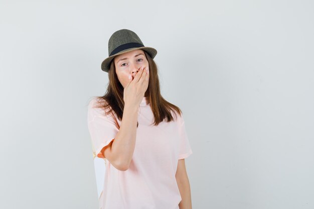 Молодая женщина держит руку на рту в розовой футболке, шляпе и выглядит удивленным, вид спереди.
