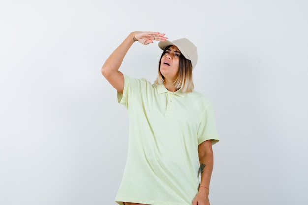 Бесплатное фото Молодая женщина держит руку возле кепки, чтобы четко видеть в футболке, кепке и смотрит сосредоточенно, вид спереди.
