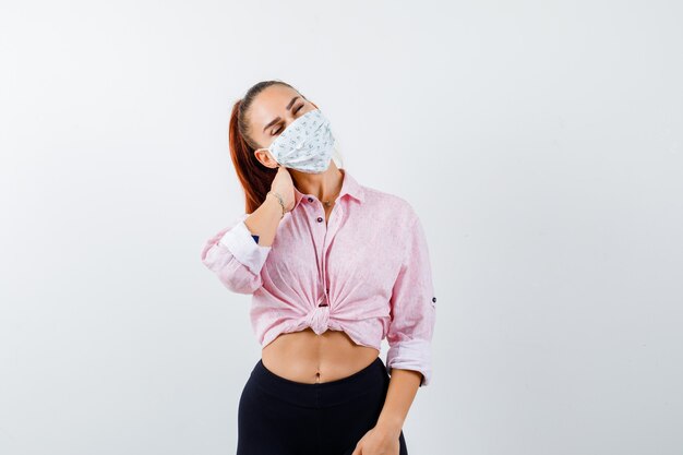 Young female holding hand on neck in shirt, pants, medical mask and looking exhausted. front view.
