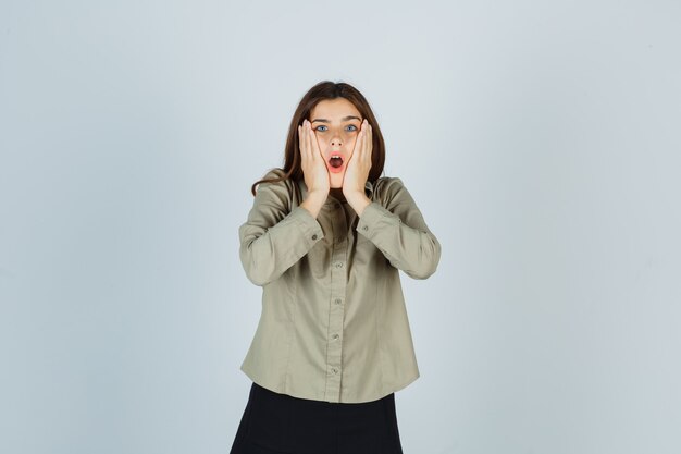 Young female holding hands on cheeks in shirt, skirt and looking shocked