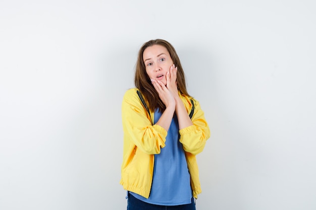 Young female holding hands on cheeks in t-shirt, jacket and looking alluring, front view.