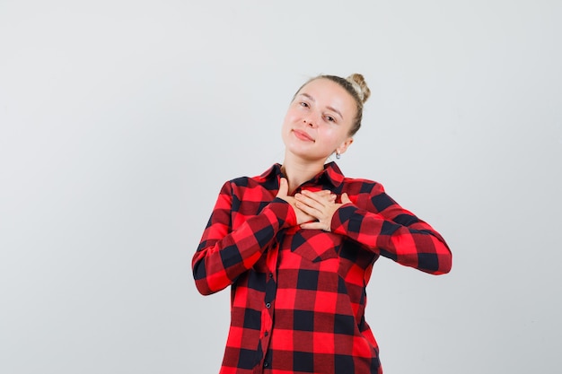 Young female holding hands on chest in checked shirt and looking cute , front view.