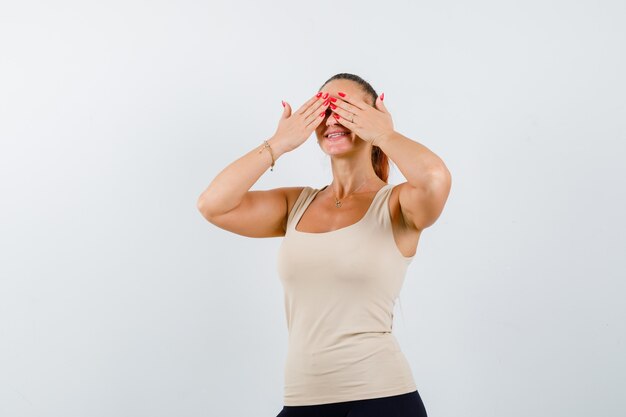 Young female holding hands on eyes in beige tank top and looking cute. front view.