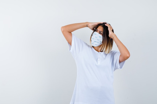 Young female holding hands on head in t-shirt, mask and looking hesitant , front view.