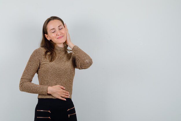 Young female holding hands on her face while closing her eyes in golden blouse and looking impressive , front view. space for text