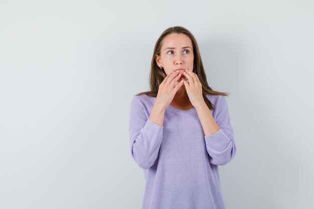 Young female holding hands on her lips while looking away in lilac blouse and looking focused 