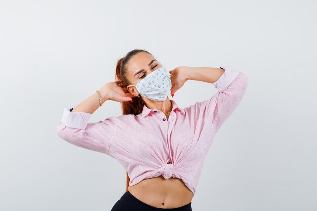 Бесплатное фото Молодая женщина, взявшись за руки в рубашке, штанах, медицинской маске и уверенно глядя, вид спереди.