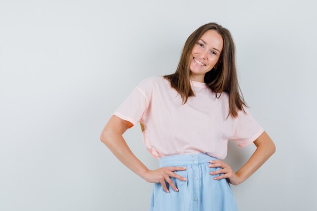 Young female holding hands on waist in t-shirt, skirt and looking cheerful. front view.