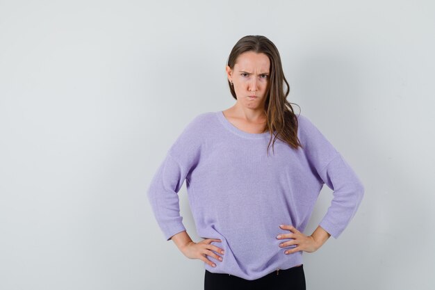 Young female holding her hands on waist in lilac blouse and looking angry. front view.