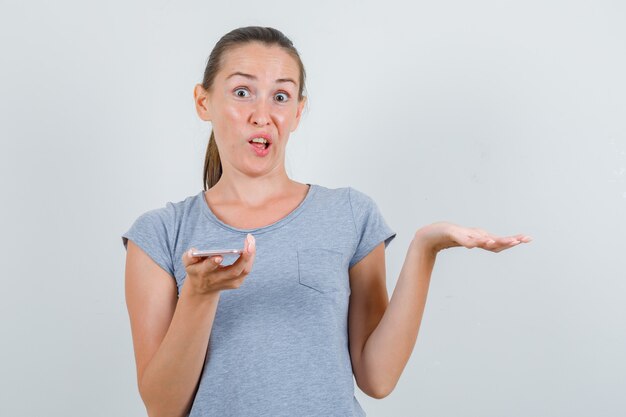 Young female holding mobile phone in grey t-shirt and looking confused. front view.