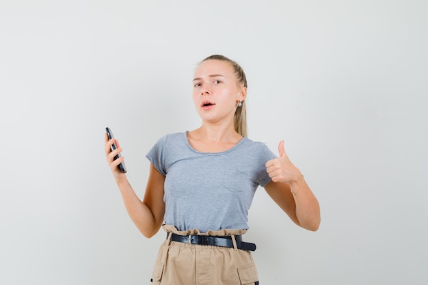 Young female holding mobile phone, showing thumb up in t-shirt, pants front view.