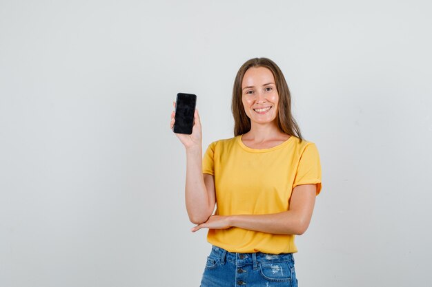 Молодая женщина держит мобильный телефон и улыбается в футболке, вид спереди шорты.
