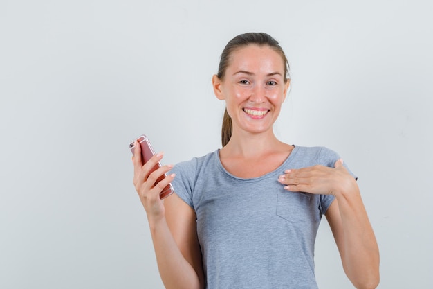 灰色のTシャツを着て、うれしそうに見える携帯電話を持っている若い女性。正面図。