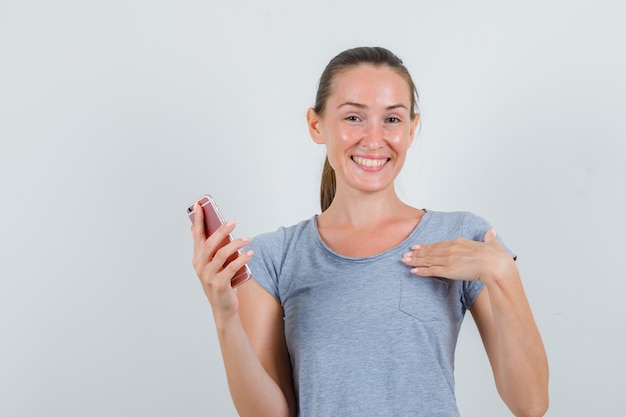 Молодая женщина держит мобильный телефон, показывая себя в серой футболке и выглядит радостной. передний план.