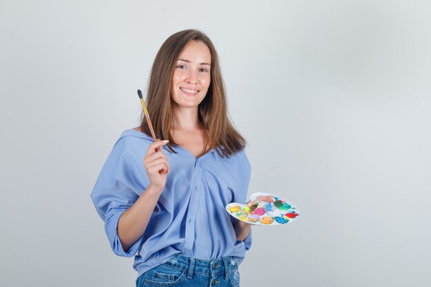 Young female holding paint brush and palette in blue shirt, shorts and looking optimistic