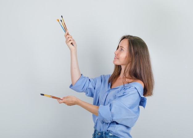 Young female holding paint brushes in shirt, shorts and looking cheerful.