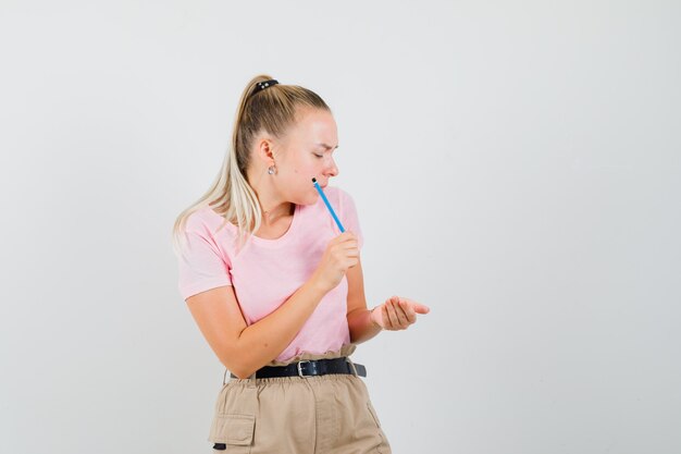 Young female holding pencil and looking at cupped hand in t-shirt, pants front view.