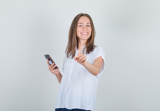 Молодая женщина держит смартфон и жестикулирует пальцем в белой рубашке
