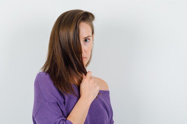 Young female holding strand of hair in violet shirt , front view.