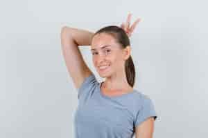 Free photo young female holding v-sign behind head as horns in grey t-shirt and looking funny , front view.