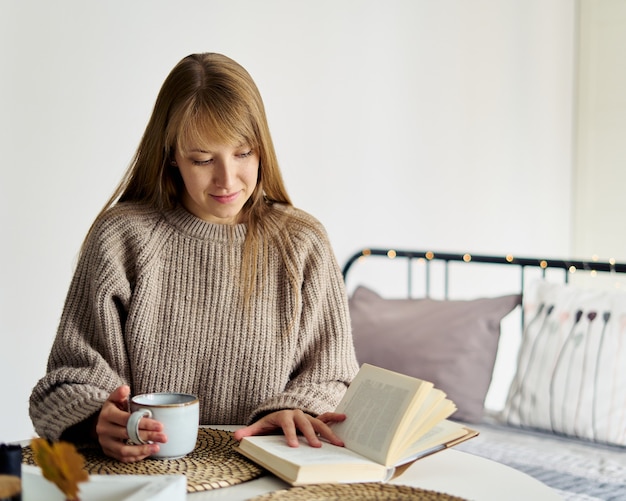스웨터를 입은 젊은 여성, 침실에서 따뜻한 차 한잔과 함께 책을 읽는 느린 삶