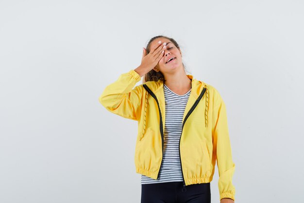 Young female in jacket, t-shirt holding hand on eye and looking happy , front view.