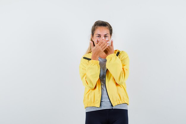 Young female in jacket, t-shirt holding hands on mouth and looking jolly , front view.