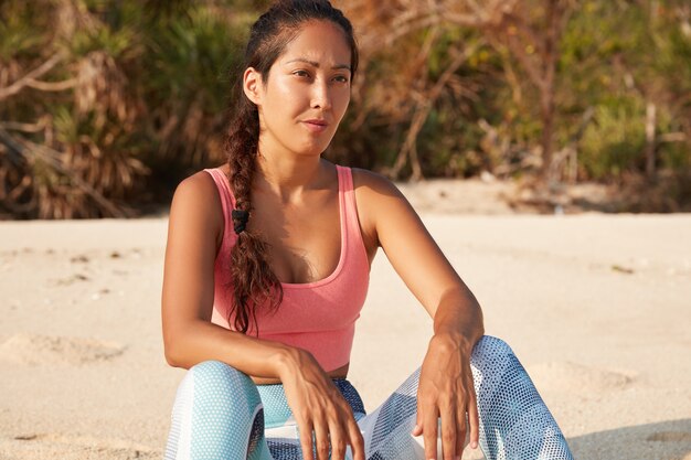 Young female jogger in active wear feels healthy, looks thoughtfully into distance, poses on sandy beach