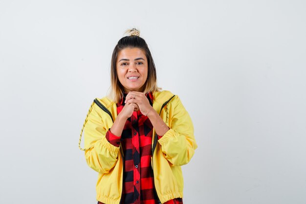 Young female keeping clasped hands near face in checkered shirt, jacket  and looking blissful , front view.