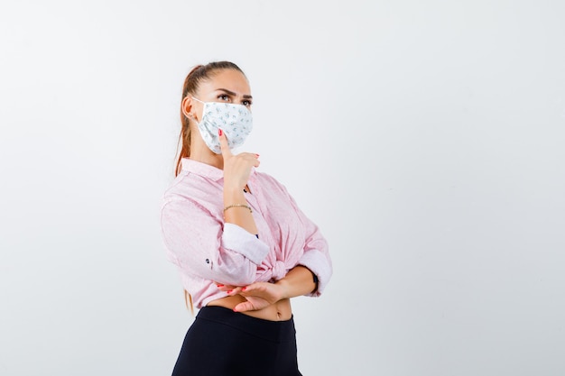 Young female keeping finger on cheek in shirt, pants, mask and looking pensive , front view.