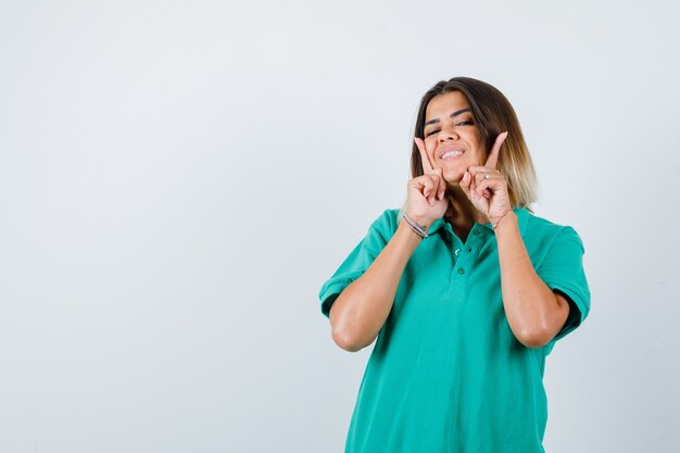 Молодая женщина держит пальцы на щеках в футболке поло и выглядит весело, вид спереди.
