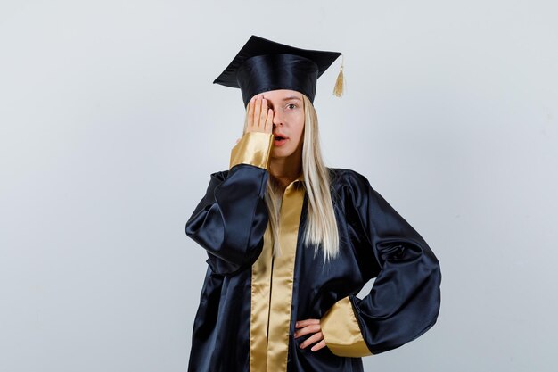 Young female keeping hand on eye in graduate uniform and looking focused.