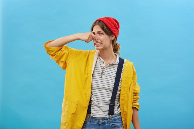 Young female keeping hand on nose having disgusting look while smelling something unpleasant isolated over blue wall. Displeased female showing her reaction on stinking smell from kitchen