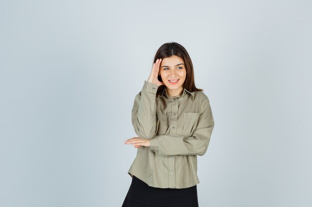 Young female keeping hand on the side of face in shirt, skirt and looking happy