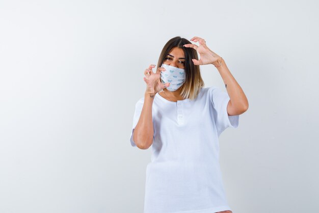 Young female keeping hands in aggressive manner in t-shirt, mask and looking stressed , front view.