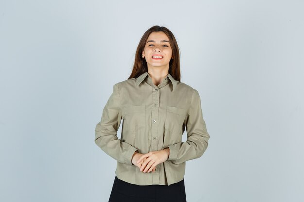 Young female keeping hands in front of her in shirt, skirt and looking merry