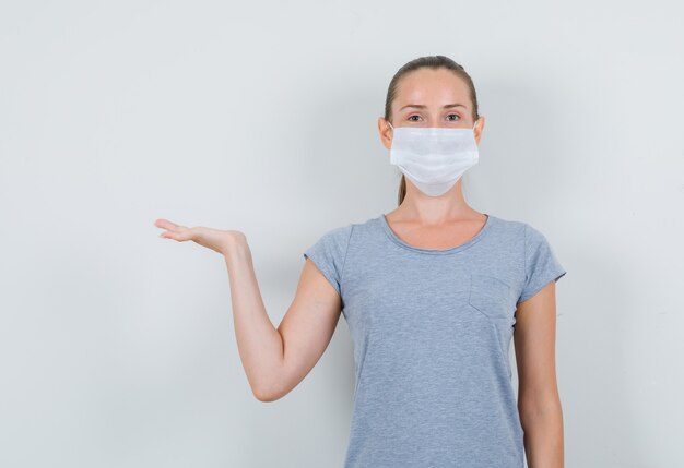 Young female keeping palm as holding something in grey t-shirt, mask front view.