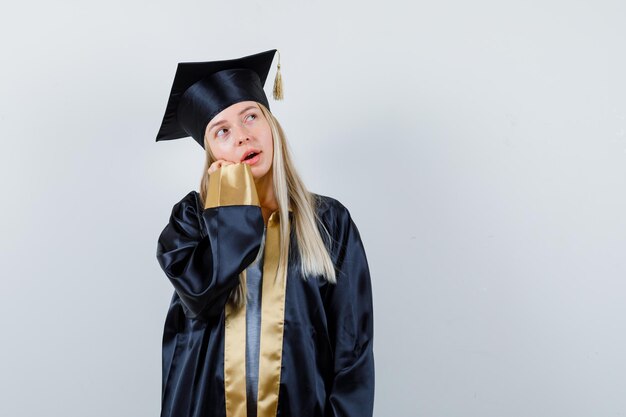 大学院の制服を着て、夢のように見える若い女性の頬を手に傾けます。