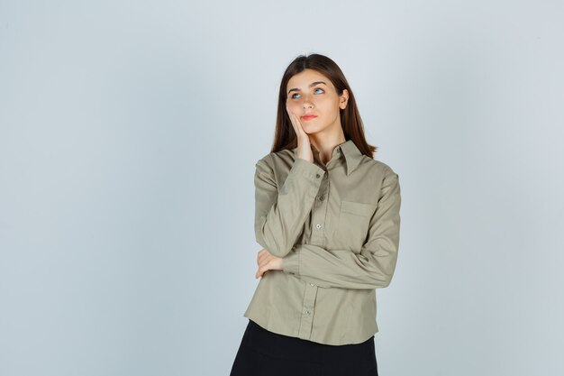 Young female leaning cheek on hand in shirt, skirt and looking pensive , front view.