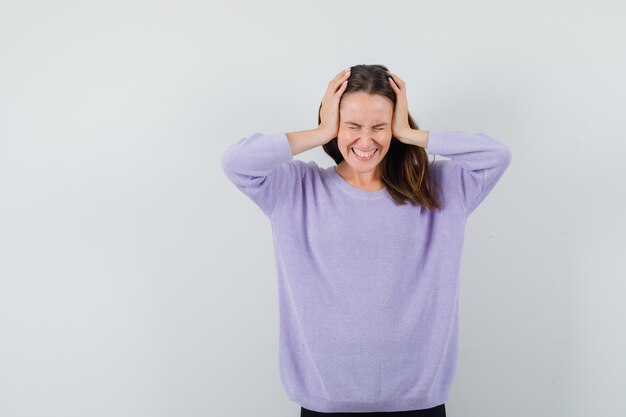 Young female in lilac blouse clasping head with hands and looking joyful 