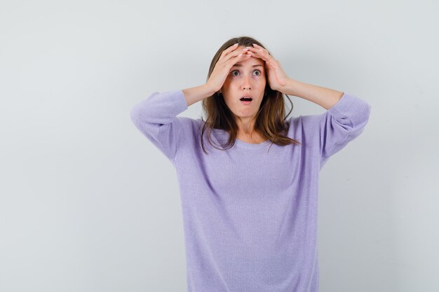 Young female in lilac blouse clasping head with hands and looking scared 