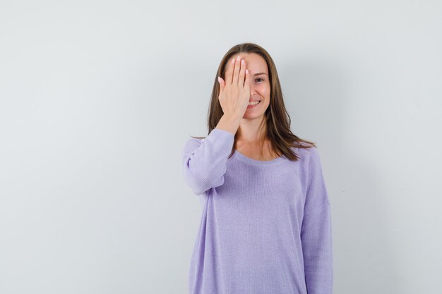 Young female in lilac blouse covering her half face with her hand and looking joyful 