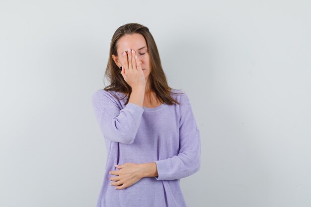 Young female in lilac blouse holding hand on her face and looking bored 